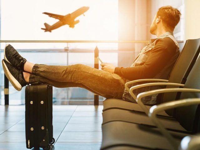 man siting in airport terminal with wheeled carry-on backpack